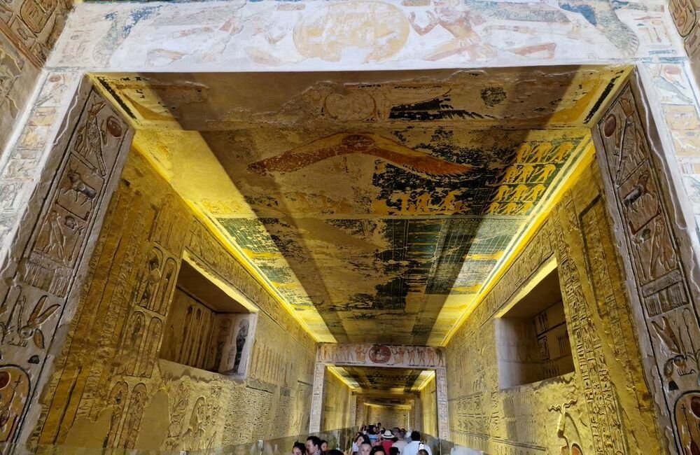 KV6 (Tomb of Ramesses IX) featuring carved hieroglyphic walls, painted astronomical ceiling scenes, and tourists walking down the passageway.